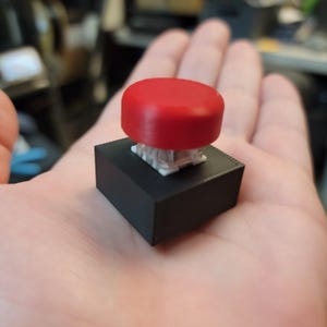 May include: A red button with a rounded top sits atop a black rectangular base. The button is held in a person's hand. The button's base has a white, intricate mechanism visible beneath the red top.