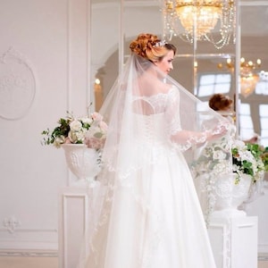May include: A bride wearing a white wedding dress with lace detailing and a long veil. The dress has a sweetheart neckline and three-quarter sleeves. The bride is standing in a white room with a chandelier in the background.