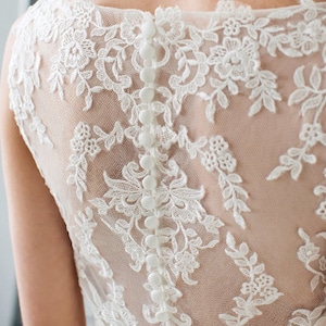 以下が含まれることがあります： 背中がボタンで留められた白いレースのウェディングドレスです。