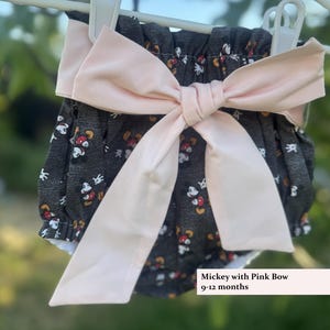 May include: Dark gray baby bloomers with a pattern of Mickey Mouse faces in red, white, and yellow. A large, light pink bow is attached to the front. The text "Mickey with Pink Bow 9-12 months" is visible.