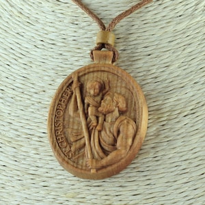 Puede incluir: Un collar con un colgante de madera tallado con la imagen de San Cristóbal llevando a un niño. El colgante es de forma ovalada y tiene las palabras "Saint Christopher" inscritas alrededor del borde.