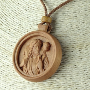 Può includere: Una collana con un ciondolo in legno intagliato con l'immagine di Gesù che porta un agnello sulle spalle. Il ciondolo è rotondo e ha un cordino in pelle marrone.
