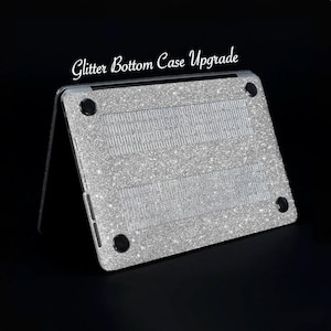 Puede incluir: Una funda para portátil plateada con la parte inferior de purpurina. La funda tiene un acabado de purpurina plateada y está diseñada para adaptarse a un portátil.