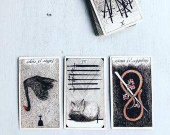 以下が含まれることがあります： 白黒のイラストが描かれたタロットカード3枚。最初のカードは赤いクチバシの黒い白鳥、2枚目は8本の剣の上に寝ている猫、3枚目は剣が突き刺さったヘビが描かれている。