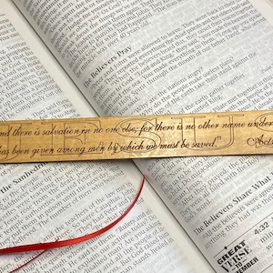 May include: A wooden bookmark with a red ribbon. The bookmark features a light brown wood background with the words "And there is salvation in no one else; for there is no other name under heaven that has been given among men by which we must be saved."  The text is in a dark brown font and the words "JESUS" are in a larger font. The bookmark also includes the scripture reference "Acts 4:12".
