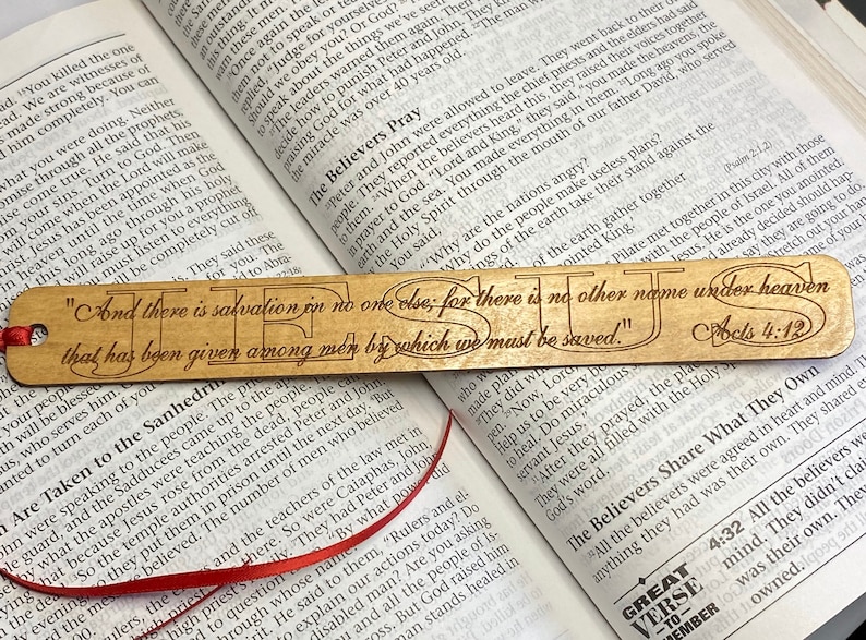 May include: A wooden bookmark with a red ribbon. The bookmark features a light brown wood background with the words "And there is salvation in no one else; for there is no other name under heaven that has been given among men by which we must be saved."  The text is in a dark brown font and the words "JESUS" are in a larger font. The bookmark also includes the scripture reference "Acts 4:12".