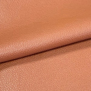 May include: Close-up of a textured, peach-colored leather fabric. The fabric is folded in half, showing the texture and color.