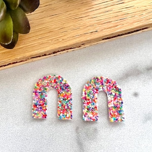 Puede incluir: Un par de pendientes de arco de purpurina arcoíris. Los pendientes están hechos de acrílico transparente y están cubiertos de purpurina de colores.