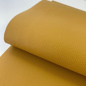 May include: A close-up of a piece of mustard yellow faux leather fabric. The fabric has a textured surface that resembles leather.