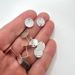 May include: Silver earring blanks with a textured finish. The blanks are round and have a post on the back. There are eight blanks in the image.