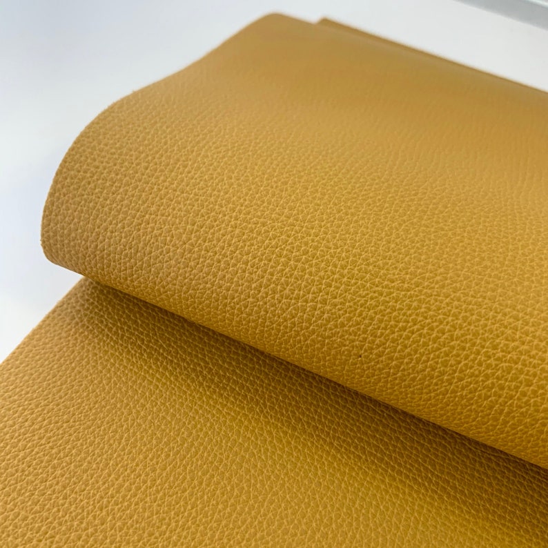 May include: A close-up of a piece of mustard yellow faux leather fabric. The fabric has a textured surface that resembles leather.