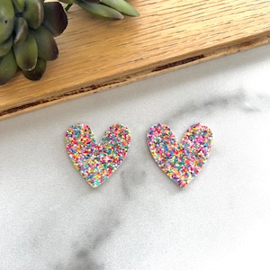 May include: Two colorful glitter heart-shaped earrings. The hearts are a light pink color and covered in a variety of multi-colored glitter.