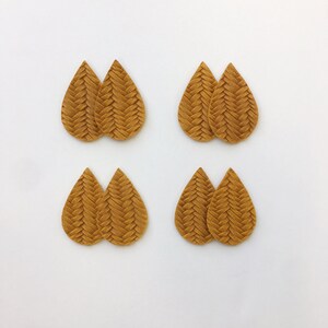 May include: Four pairs of teardrop-shaped earrings in a warm, golden-brown hue. Each earring has a woven, textured design, creating a braided appearance. The earrings are arranged on a white background, showcasing their intricate details.