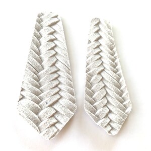 Puede incluir: Un par de pendientes de cuero blanco con un dise&ntilde;o trenzado. Los pendientes son largos y estrechos, con una punta puntiaguda.