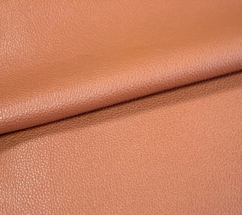 May include: Close-up of a textured, peach-colored leather fabric. The fabric is folded in half, showing the texture and color.