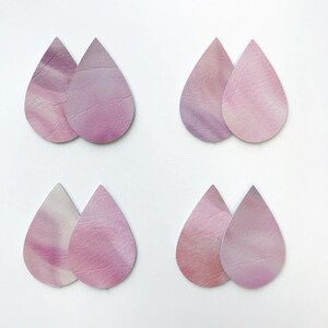 May include: Eight teardrop-shaped leather earring cutouts in pink and purple hues. The leather has a marbled or tie-dye effect, with variations in colour intensity. The cutouts are arranged in pairs against a white background, suitable for crafting.