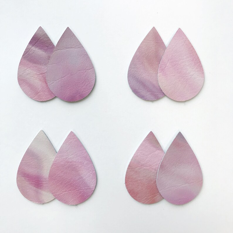 May include: Eight teardrop-shaped leather earring cutouts in pink and purple hues. The leather has a marbled or tie-dye effect, with variations in colour intensity. The cutouts are arranged in pairs against a white background, suitable for crafting.
