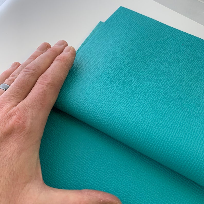 May include: A close-up of a teal-colored leather fabric. The fabric has a textured surface and is folded in half.