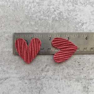 Może przedstawiać: Dwa czerwone sk&oacute;rzane kształty serc umieszczone na linijce. Serca mają około 3,8 cm szerokości i 4,4 cm wysokości.
