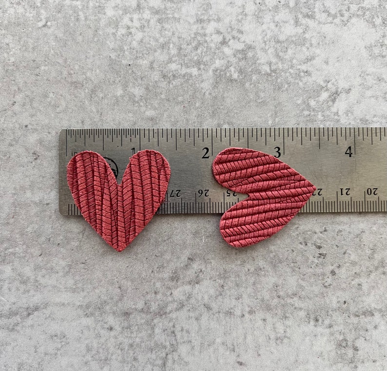 Może przedstawiać: Dwa czerwone sk&oacute;rzane kształty serc umieszczone na linijce. Serca mają około 3,8 cm szerokości i 4,4 cm wysokości.