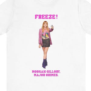 May include: White t-shirt featuring the pink text "FREEZE!" and "MORGAN GILLORY. MAJOR CRIMES." The shirt has a graphic of a person wearing a pink jacket, black skirt, and blue boots.