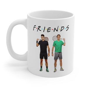 May include: White ceramic mug with a black and white image of two tennis players holding rackets. The text "FRIENDS" is printed in black with colorful dots above the letters.