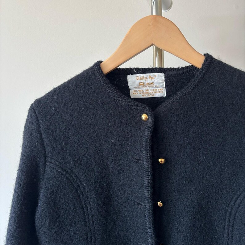 Puede incluir: Un c&aacute;rdigan de lana negro con botones dorados. La chaqueta vintage tiene un cuello redondo y est&aacute; colgada de una percha de madera. La etiqueta dice "Tallo-ho".