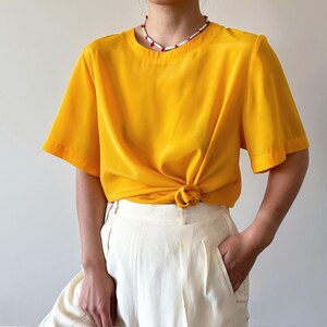 Puede incluir: Una blusa de manga corta de color amarillo brillante con cuello redondo. La blusa est&aacute; anudada a la cintura y se lleva con pantalones blancos.