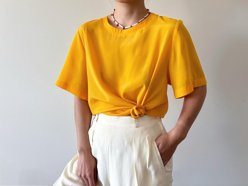 Puede incluir: Una blusa de manga corta de color amarillo brillante con cuello redondo. La blusa est&aacute; anudada a la cintura y se lleva con pantalones blancos.