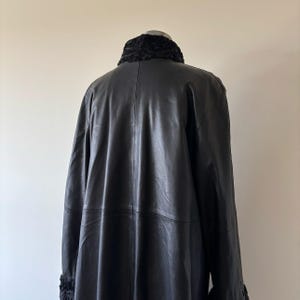 May include: A black leather coat featuring a smooth finish and a textured black fur collar and matching fur cuffs. The coat is displayed on a mannequin, showing a long-line silhouette with visible vertical seam detailing along the back panel.