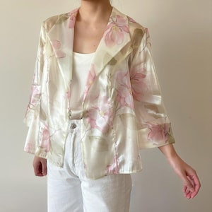 May include: A sheer, ivory-colored jacket with a floral print in shades of pink. The jacket has a shawl collar and a loose, flowing fit.