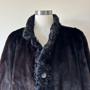 May include: A dark brown coat featuring a textured, curly black trim along the collar and front placket. The garment includes a visible round button closure on the front, showcasing a contrast between the smooth main body and the curly texture.