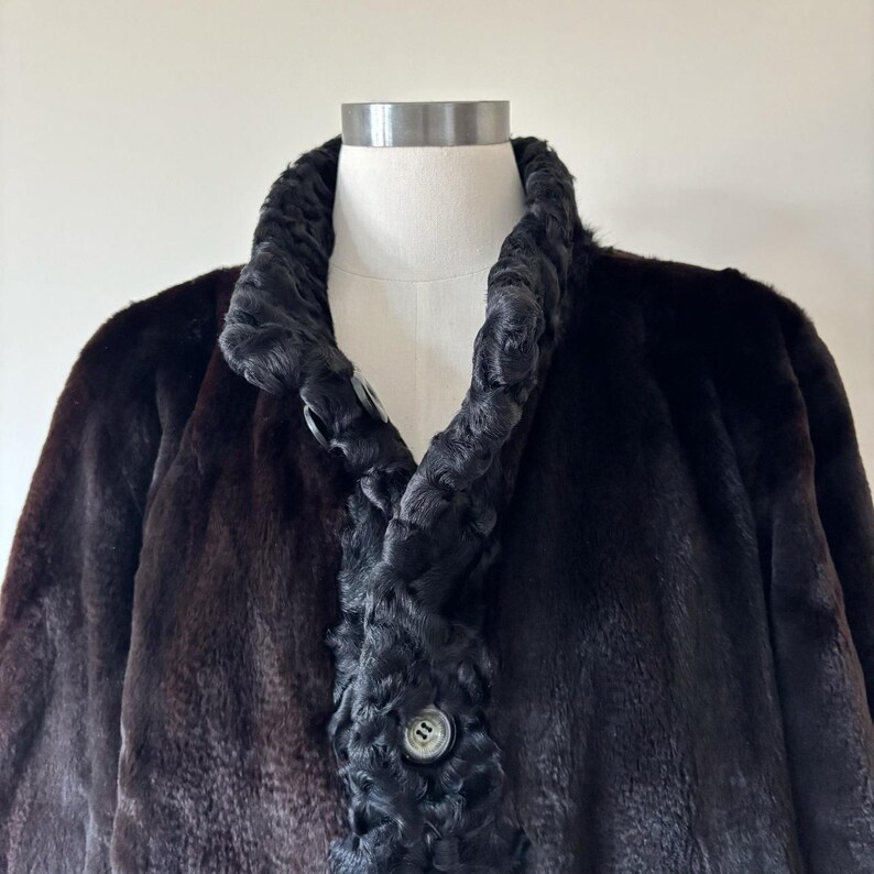 May include: A dark brown coat featuring a textured, curly black trim along the collar and front placket. The garment includes a visible round button closure on the front, showcasing a contrast between the smooth main body and the curly texture.