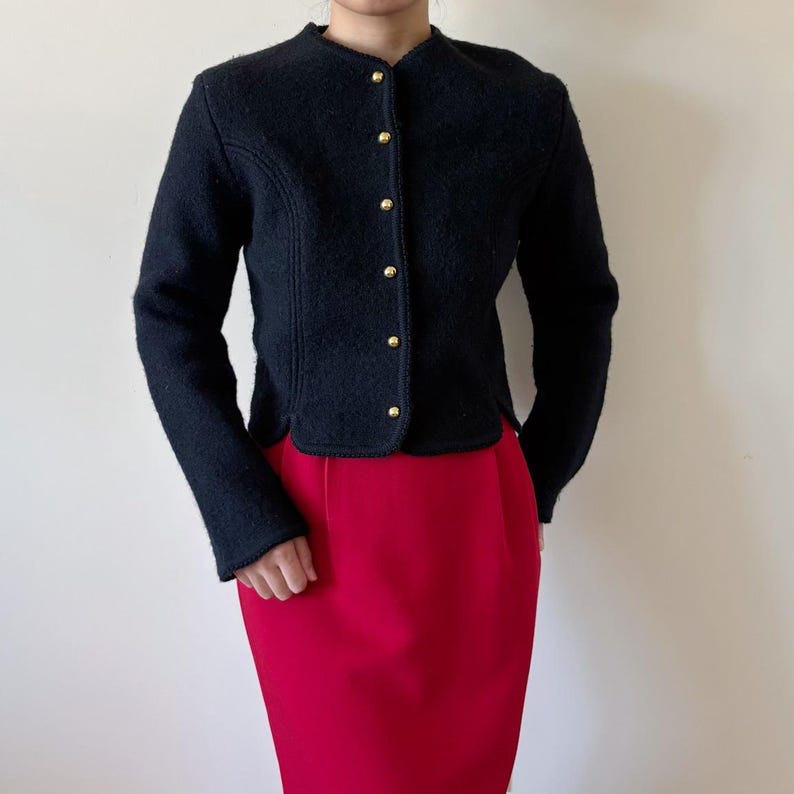 Puede incluir: Chaqueta negra de lana con cuello redondo y botones dorados. La chaqueta se combina con una falda l&aacute;piz roja. La chaqueta tiene mangas largas y un corte entallado. El atuendo es adecuado para un entorno profesional o formal.