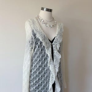 May include: A cream-colored, open-front cardigan with a delicate, open-weave design. The cardigan features a ruffled trim along the front and sleeves. A silver necklace with floral accents is also visible.