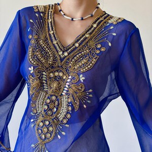 May include: A sheer blue top with long sleeves and a V-neckline. The top is embellished with gold and white beading in a floral design.