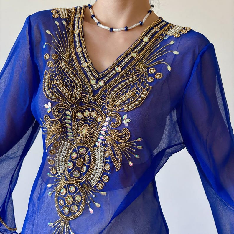 May include: A sheer blue top with long sleeves and a V-neckline. The top is embellished with gold and white beading in a floral design.