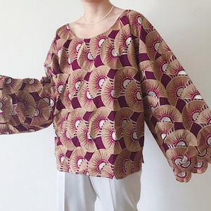 May include: A long-sleeved blouse with a scoop neckline. The fabric features a repeating fan-like pattern in burgundy, tan, and silver. The sleeves have a scalloped edge. The blouse is a loose fit.