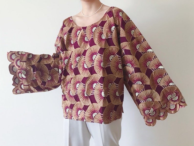 May include: A long-sleeved blouse with a scoop neckline. The fabric features a repeating fan-like pattern in burgundy, tan, and silver. The sleeves have a scalloped edge. The blouse is a loose fit.