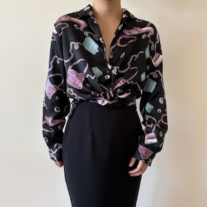 May include: Black long-sleeve blouse with a repeating pattern of purses, chains, and other accessories in shades of purple, blue, and white. The blouse is knotted at the waist and paired with a black skirt. The blouse has a collared neckline and button-down front.