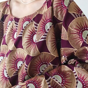 May include: A maroon-colored top with a repeating embroidered fan pattern in gold, red, and white. The top has a scoop neckline and long sleeves. A delicate pearl necklace is visible.