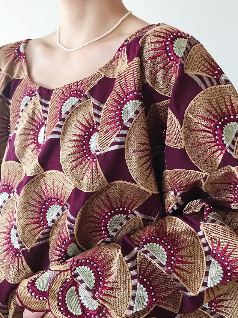May include: A maroon-colored top with a repeating embroidered fan pattern in gold, red, and white. The top has a scoop neckline and long sleeves. A delicate pearl necklace is visible.