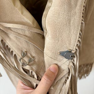 May include: Close-up of a tan suede jacket with fringe detailing. The jacket features a metal clasp closure and decorative fringe along the edges. The interior lining is a lighter tan color.