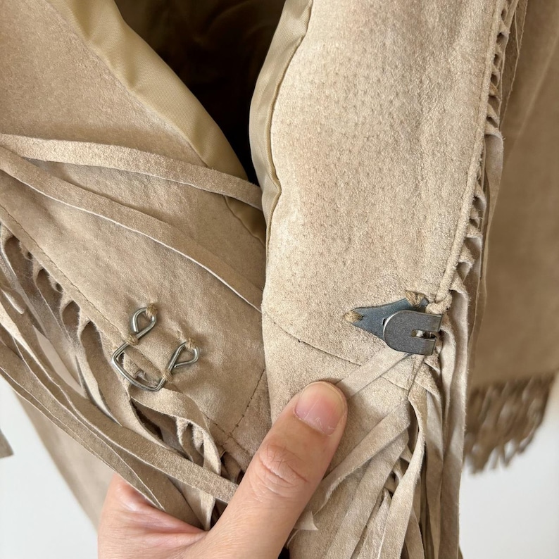 May include: Close-up of a tan suede jacket with fringe detailing. The jacket features a metal clasp closure and decorative fringe along the edges. The interior lining is a lighter tan color.