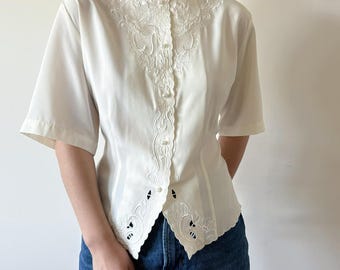 Puede incluir: Una blusa blanca con mangas cortas y cuello festoneado. La blusa presenta intrincados detalles bordados alrededor del cuello y en la parte delantera, con pequeños botones y diseños recortados en el dobladillo. La blusa se combina con unos vaqueros azules.