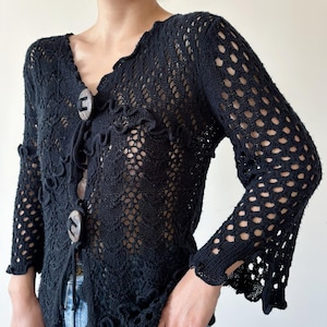 May include: Black crochet cardigan with a V-neck and long sleeves. The cardigan has a delicate openwork pattern and features two wooden buttons.