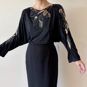 May include: Black dress with long sleeves and a knee-length skirt. The top is decorated with gold and iridescent sequins in a leaf-like pattern around the neckline and on the sleeves. The dress has a simple, elegant design.