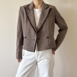 Può includere: Un blazer corto con motivo pied-de-poule marrone e beige, caratterizzato da un bordo in velluto marrone scuro lungo i revers, maniche lunghe e due bottoni marrone scuro sulla chiusura frontale. Il blazer è indossato sopra una t-shirt e pantaloni bianchi.