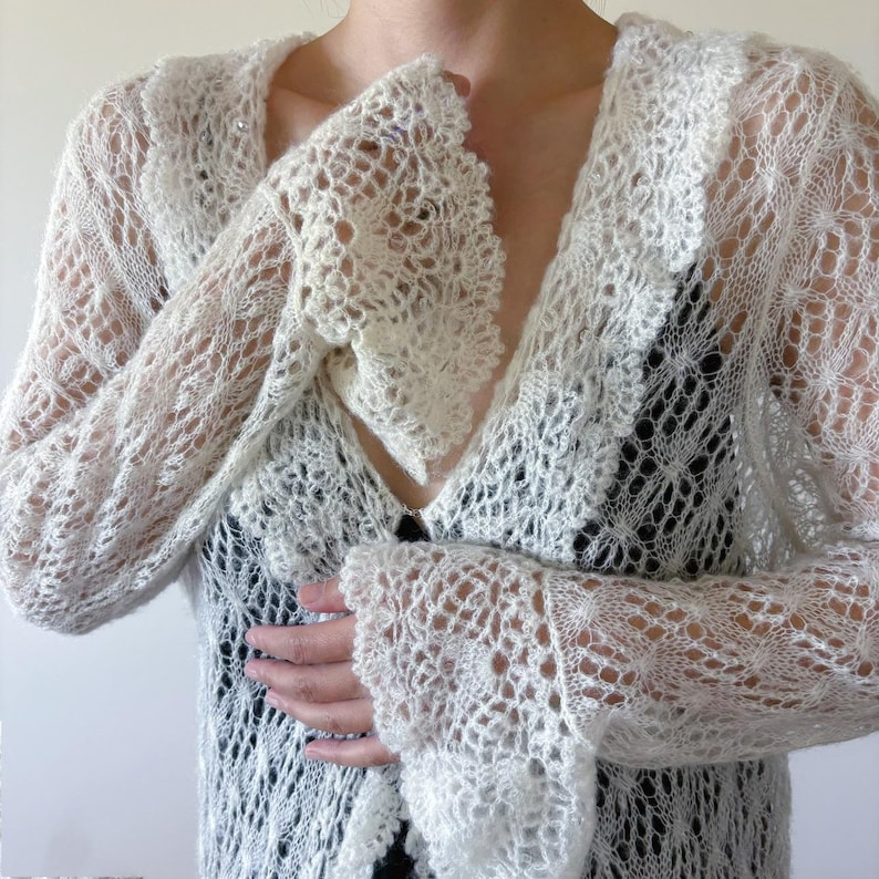 May include: Ivory-colored, open-weave cardigan with long sleeves. The delicate knit features a lacy pattern and a V-neckline. The sleeves have a flared, ruffled cuff. The cardigan is buttoned at the chest.