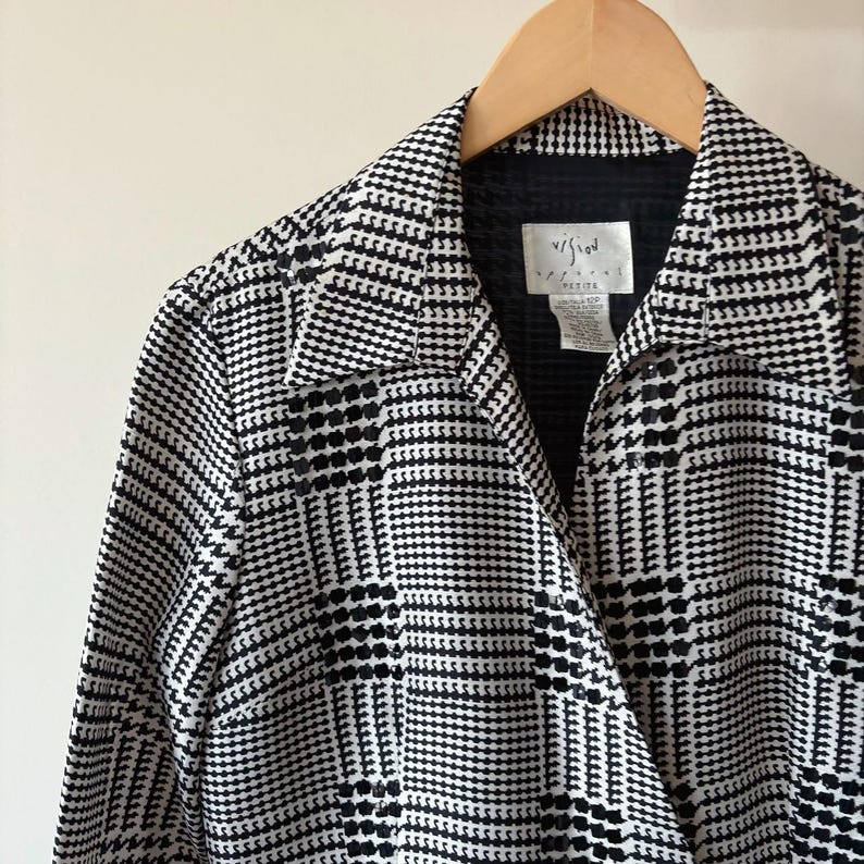 May include: A black and white plaid long-sleeved shirt with a collar and a button closure. The shirt has a 'Vision' label on the front.